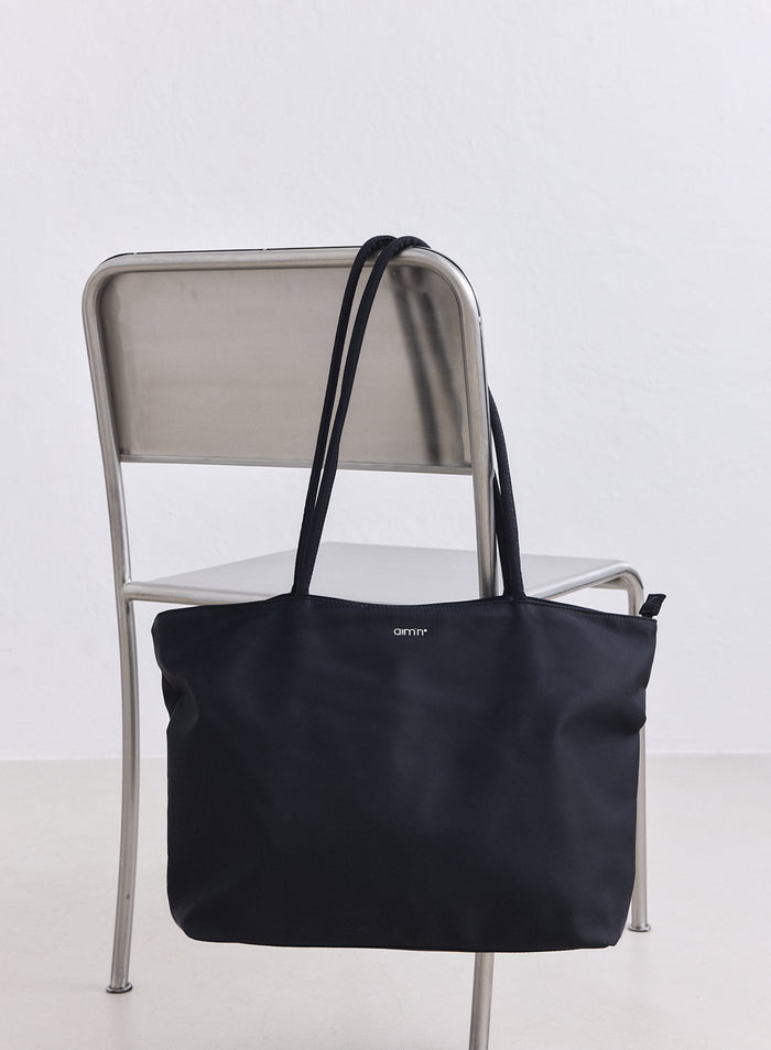 Musta nailoninen tote-laukku valkoisella aim'n-logolla roikkuu minimalistisella metallituolilla.