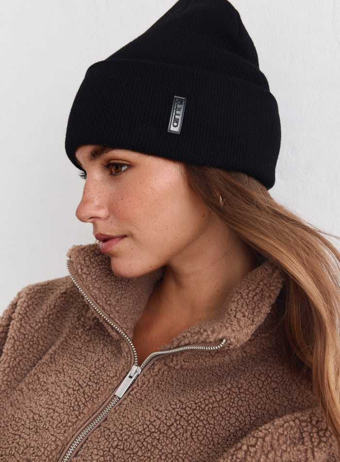 Musta neulottu aim'n Outdoor Logo Beanie yhdistettynä pehmeään ruskeaan teddyfleeletakkiin.
