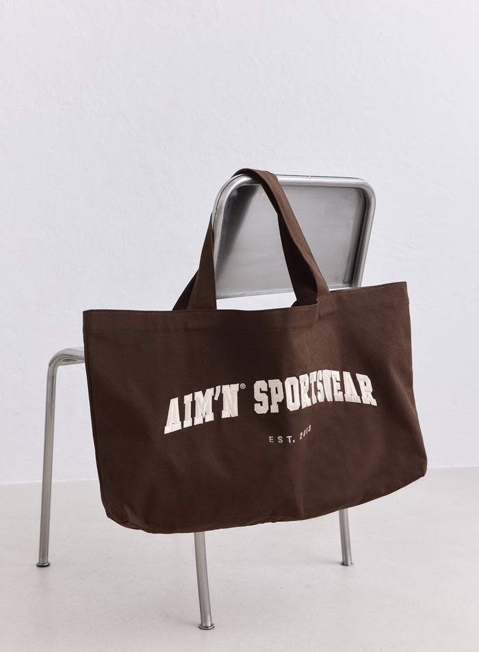 Suklaanruskea puuvillainen Varsity Tote Bag kermanvärisellä aim'n-logolla metallituolilla. [ONE-SIZE-165CM]