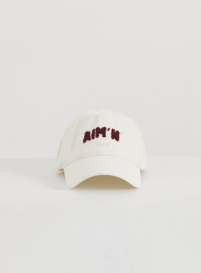 Kermavärinen puuvillainen Campus Cap tummanpunaisella pörröisellä aim'n-logolla minimalistisessa flatlay-kuvassa.