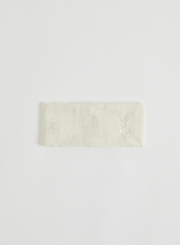 Kermanvärinen neulottu otsapanta logodetaljilla kuvattuna flat lay -asetelmassa