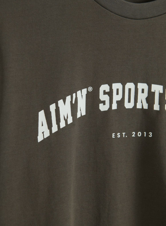 Lähikuva Dark Ivy -värisen Varsity Boxy -T-paidan ribatusta pyöreästä pääntiestä ja valkoisesta aim'n Sport Est. 2013 -grafiikasta