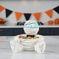 Halloween-themed mummy candy bowl with colorful candies on a table, decorated with Halloween flags in the background.