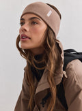 Hasselpähkinänvärinen ribbineulottu Outdoor Logo Headband valkoisella aim'n-logolla, beigen takin ja repun kera.