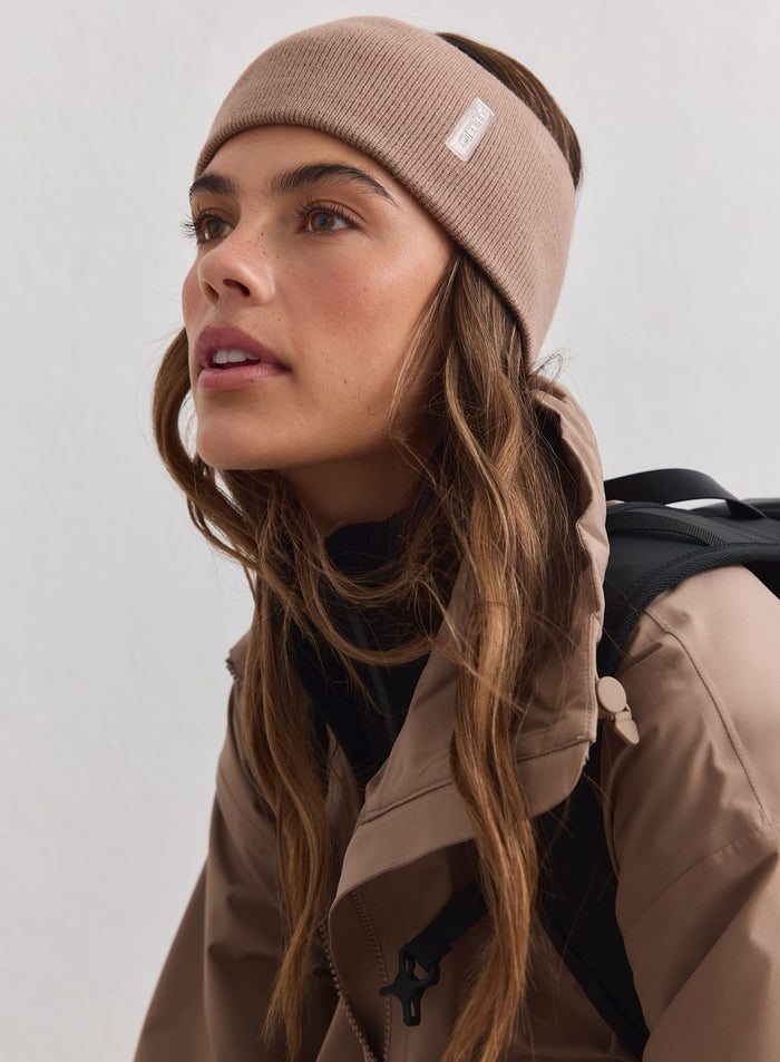 Hasselpähkinänvärinen ribbineulottu Outdoor Logo Headband valkoisella aim'n-logolla, beigen takin ja repun kera.