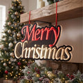 A decorative hanging Christmas sign with red and cream lettering that reads “Merry Christmas,” suspended by red ribbons in front of a fireplace with a decorated tree in the background.