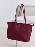 Merlot-värinen nailoninen aim'n-tote-laukku roikkuu hopeisella tuolilla minimalistisessa tilassa.