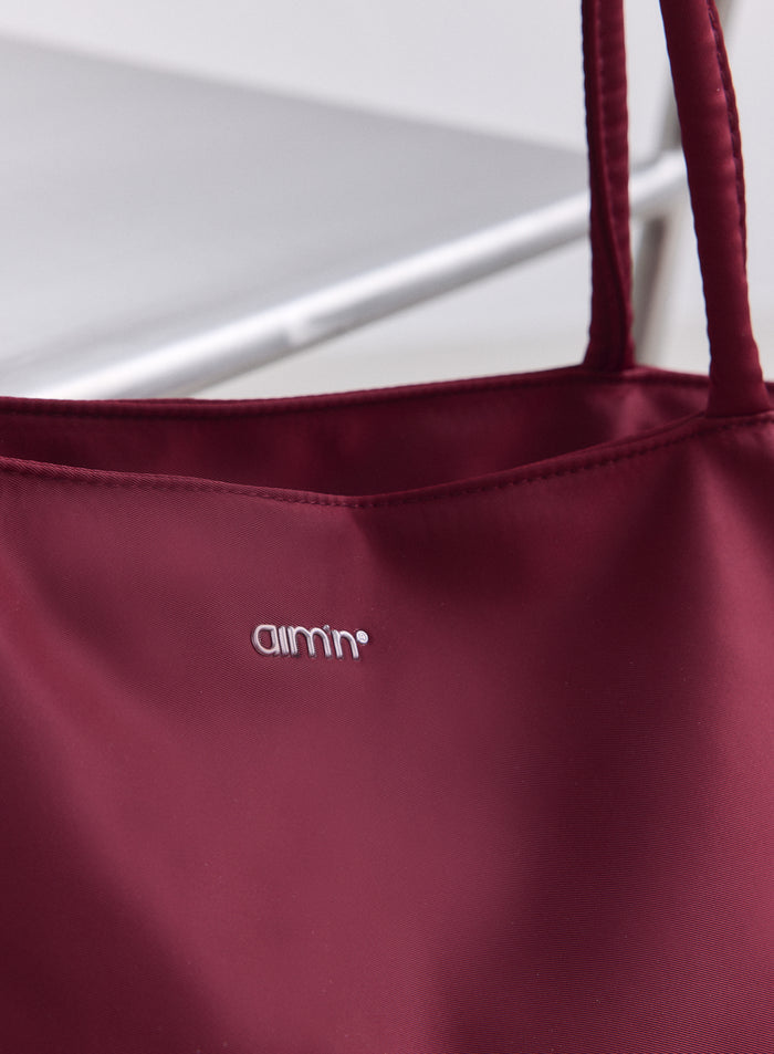 Merlot-punaisen nailonisen tote-laukun yksityiskohta, jossa on sileä pinta ja hopeinen aim'n-logo.
