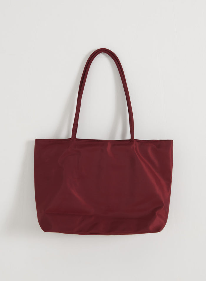 aim'n merlot-värinen nailoninen tote-laukku pitkillä olkahihnoilla ja sileällä pinnalla valkoista seinää vasten.
