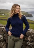 Woman wearing a navy blue cable knit sweater standing against a stone wall with a scenic background.