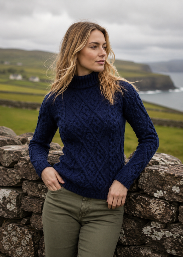Woman wearing a navy blue cable knit sweater standing against a stone wall with a scenic background.